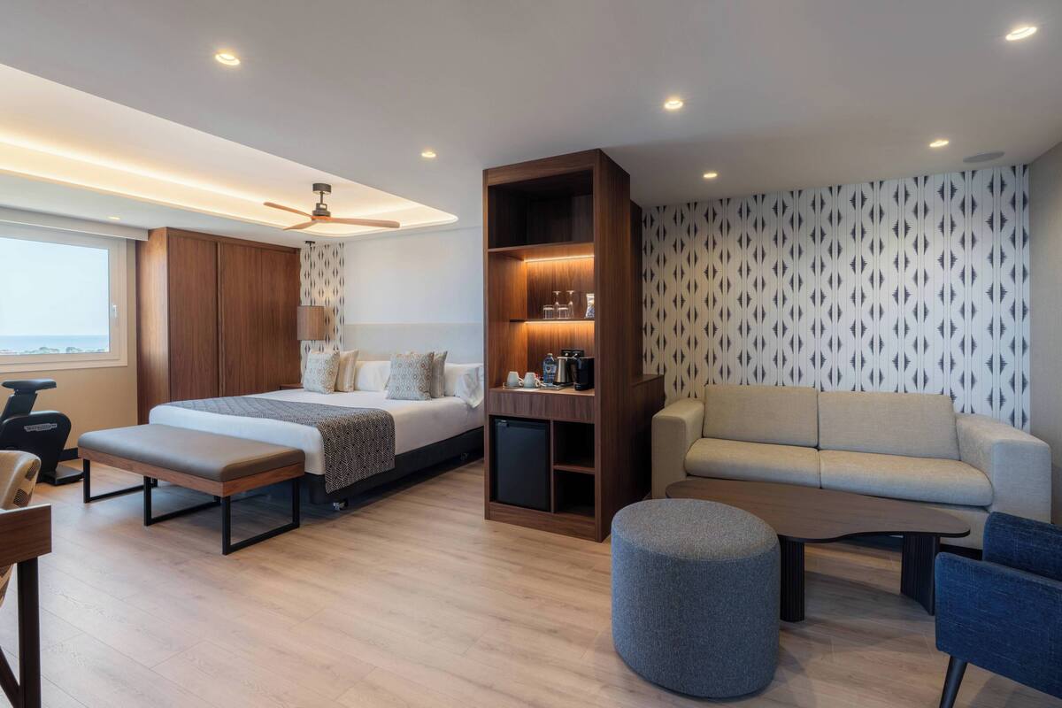 Una habitación de hotel moderna con cama, sofá, banco, reposapiés y una zona de café integrada.