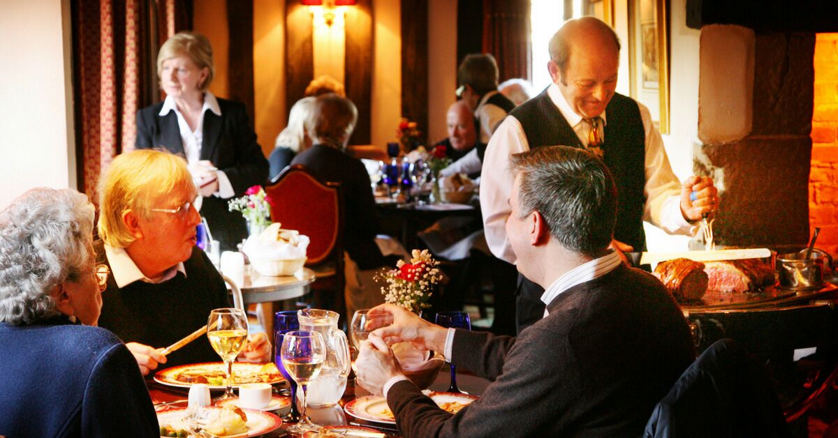 Ravintola, jossa asiakkaat istuvat pöytien ääressä, tarjoilija palvelee heitä ja taustalla näkyy tiiliseinä.