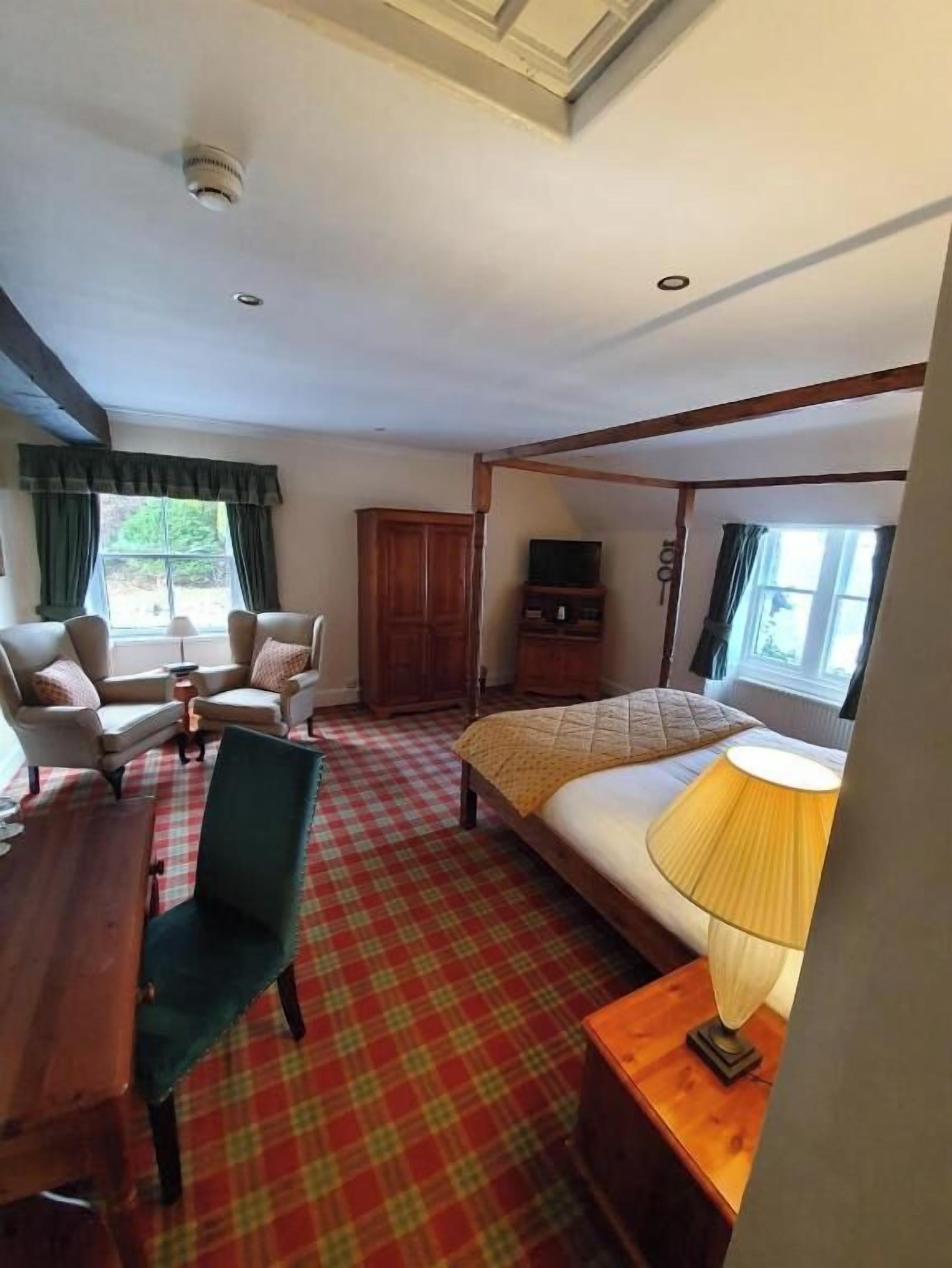 Una habitación de hotel con piso a cuadros, una cama con una lámpara amarilla, dos sillones, un armario de madera y un televisor sobre un soporte.