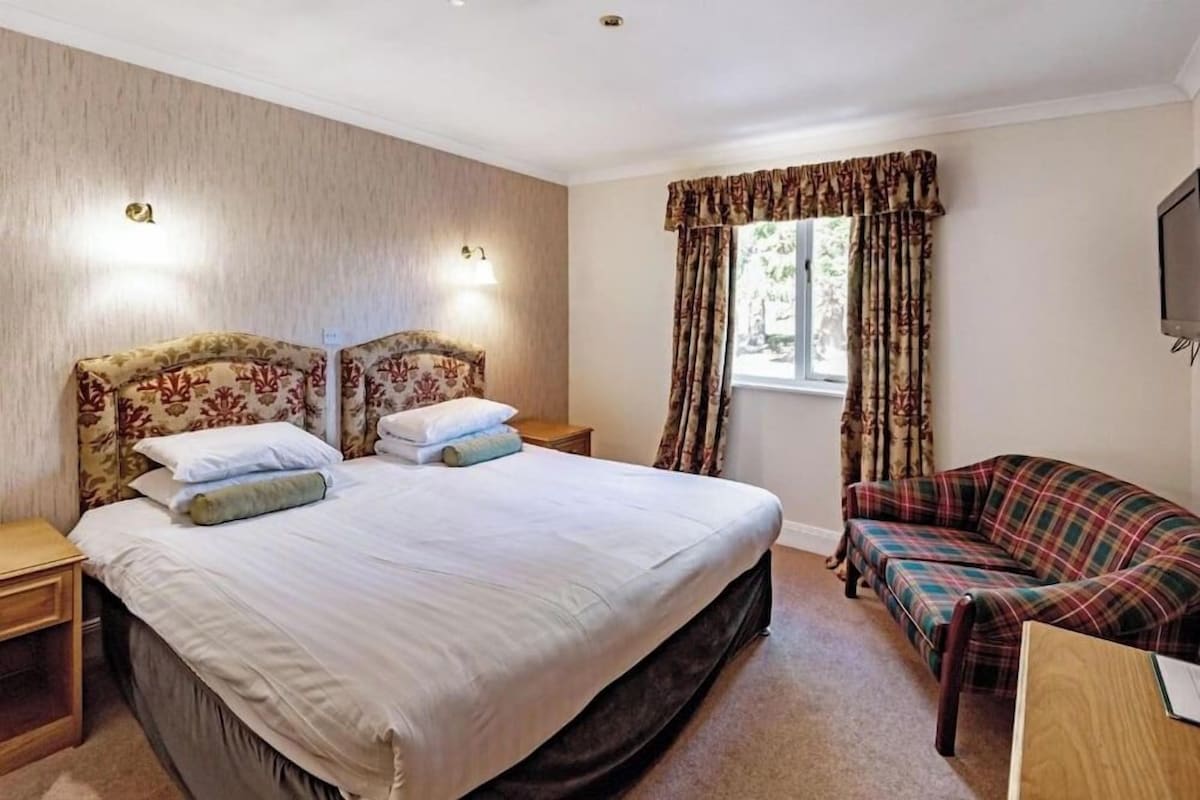 A bedroom with a large bed, two bedside tables, a flat-screen TV mounted on the wall, a window with curtains, and a plaid sofa.