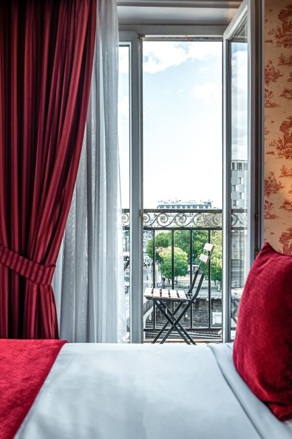 A hotel room with a bed, red curtains, and a view of a balcony with a table and chairs.