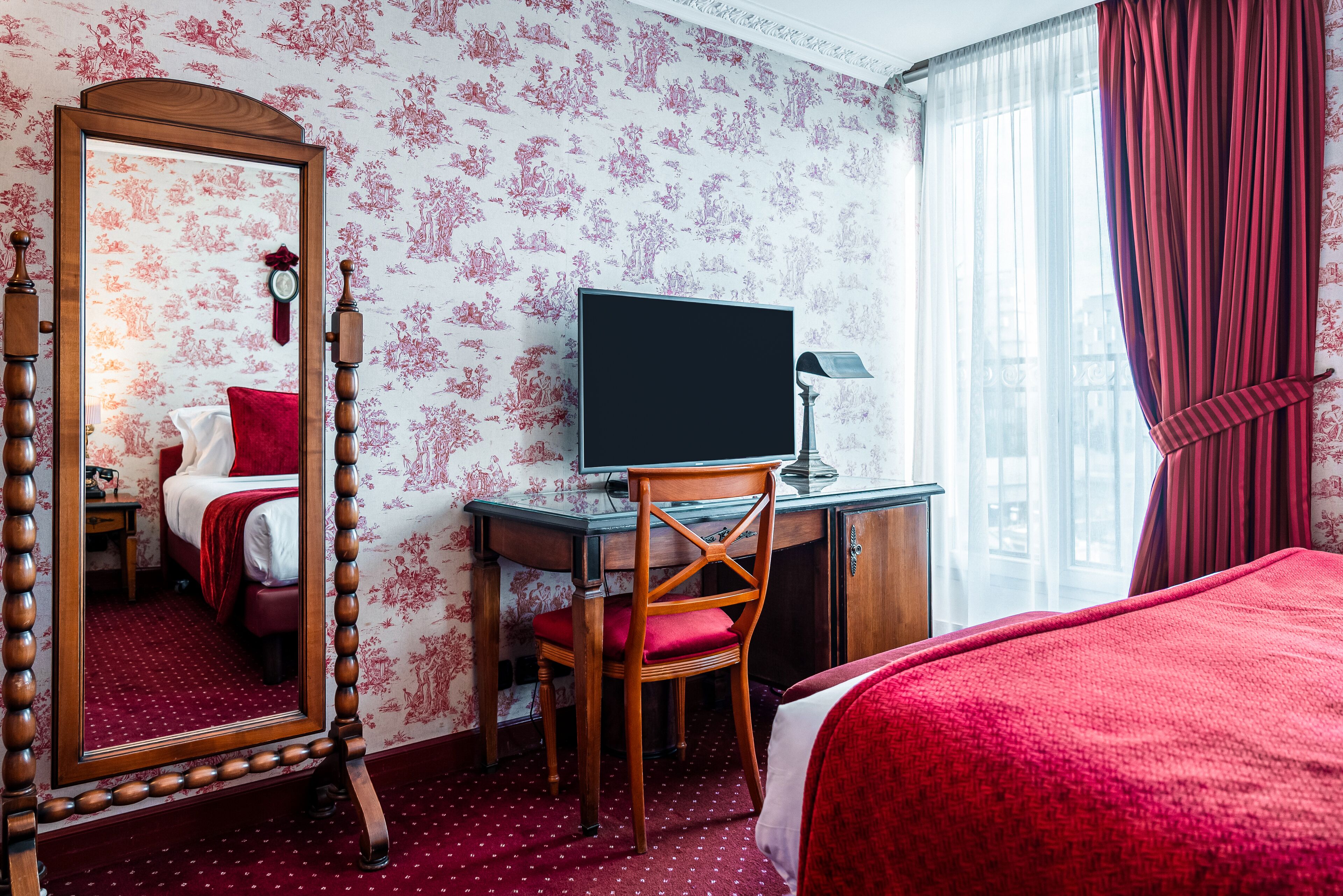 A hotel room with a large mirror, a desk with a chair, a television, and a bed with red bedding.