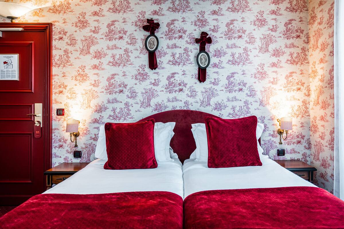 A double bed with red bedding, two wall-mounted clocks, and a floral wallpaper pattern.