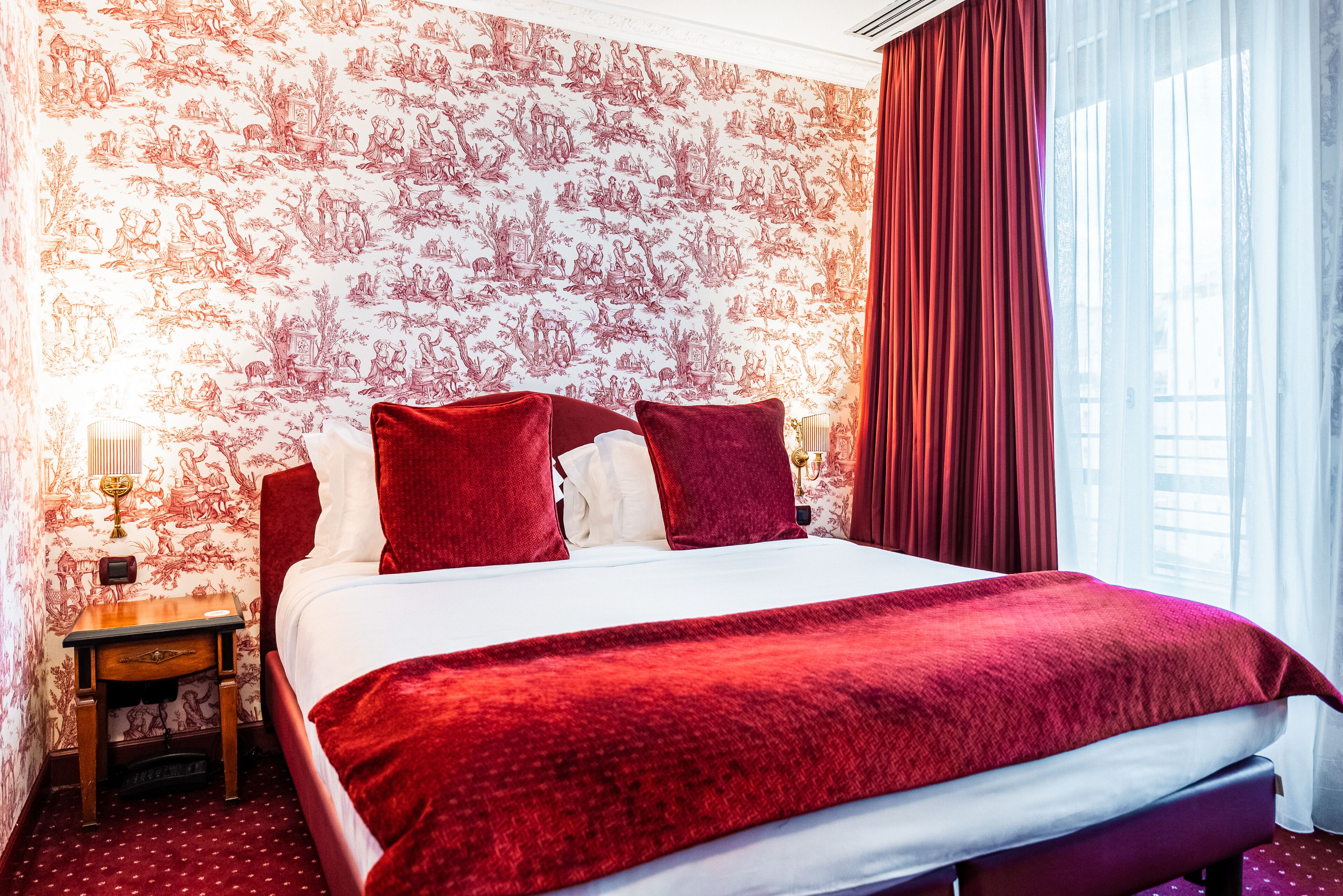 Ein Hotelzimmer mit einem großen Bett, roter Bettwäsche, einem hölzernen Nachttisch und Tapeten mit Muster.