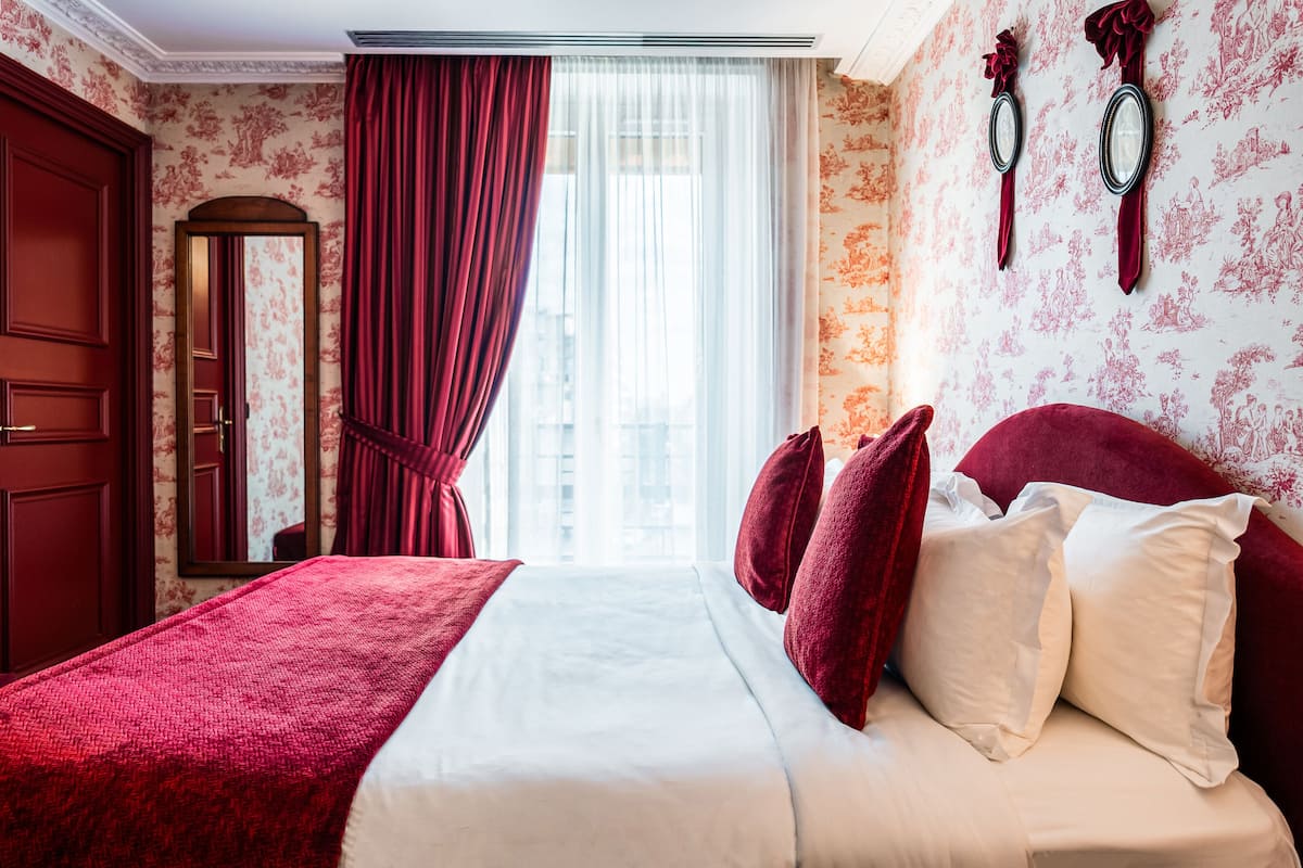 A hotel room with a large bed, red curtains, floral wallpaper, and a mirror on the wall.