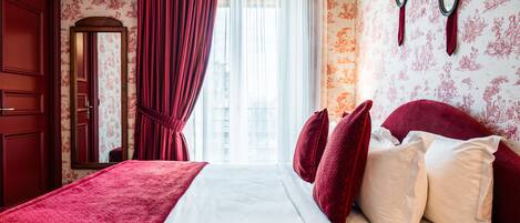 Habitación de hotel con una cama grande, cortinas rojas, papel tapiz con motivos florales y un espejo en la pared.