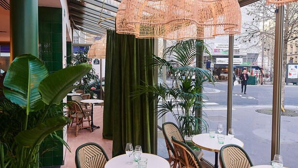 En udendørs café med fletstole og borde, grønne planter og udsigt til gaden.