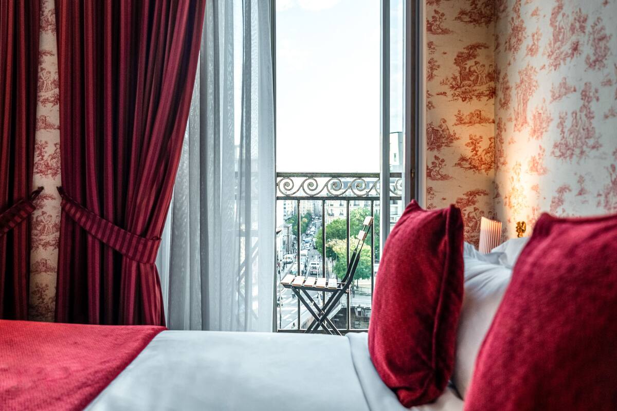 A hotel room with a balcony, a bed with red bedding, and a view of the cityscape.