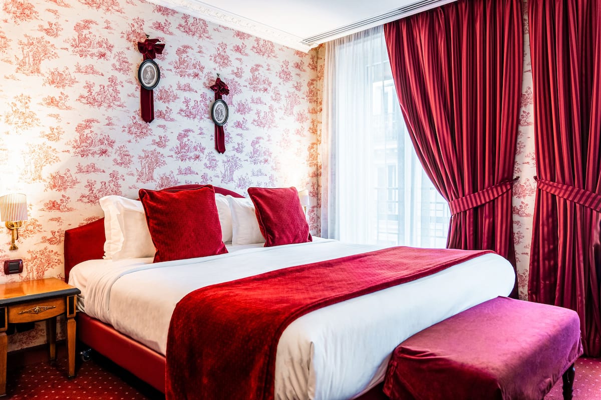 A hotel room with a large bed, red bedding, a wooden nightstand, and a wall clock.