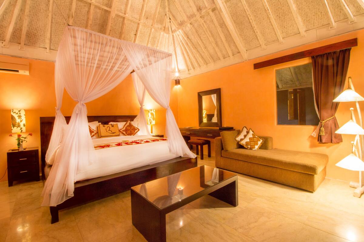 A bedroom with a four-poster bed, a sofa, a glass coffee table, and a nightstand with a lamp.