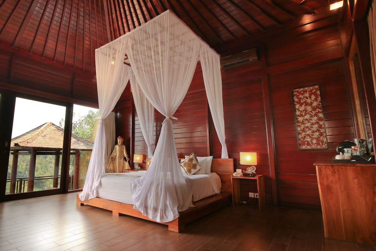 A room with a four-poster bed, wooden flooring, a large window with a view of greenery, and a nightstand with a lamp.