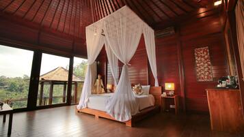 A room with a four-poster bed, wooden flooring, a large window with a view of greenery, and a nightstand with a lamp.