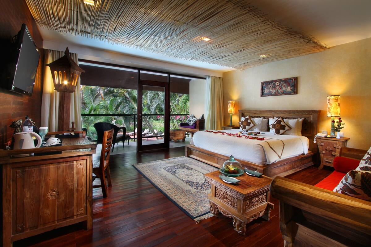 A spacious bedroom with a large bed, wooden furniture, and a view of tropical foliage through large sliding doors.