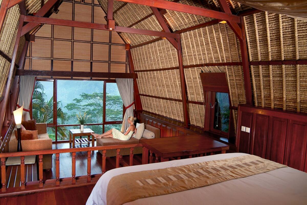 A room with a large bed, a wooden desk, and a balcony with a view of greenery.