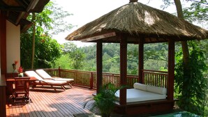 Eine Holzterrasse mit zwei Liegestühlen und einem Gazebo mit Reetdach, das auf einen Pool überblickt.
