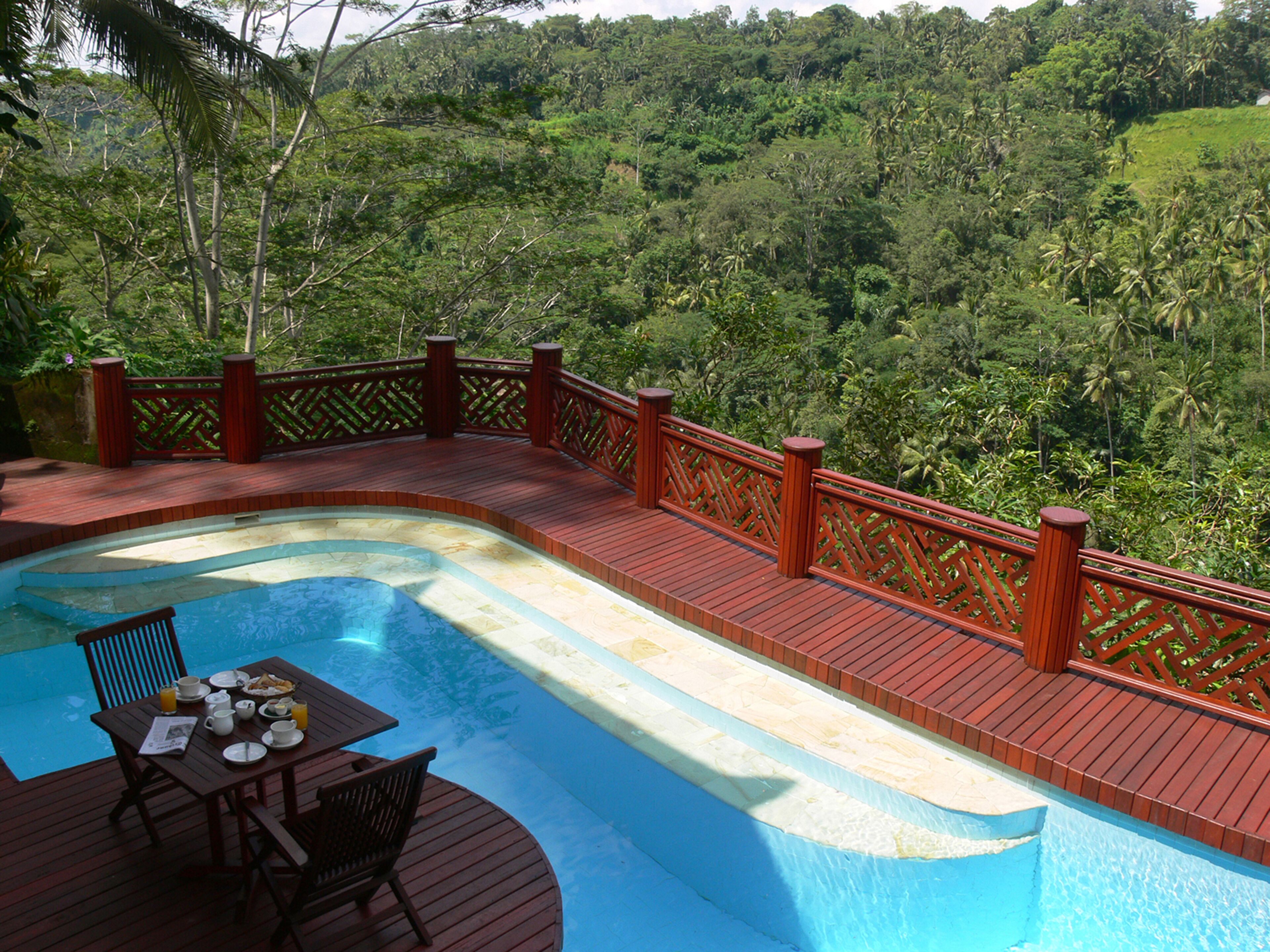 Une piscine dotée d’une terrasse en bois et d’une table avec des chaises, le tout entouré d’une végétation luxuriante.