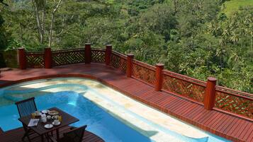 Une piscine dotée d’une terrasse en bois et d’une table avec des chaises, le tout entouré d’une végétation luxuriante.