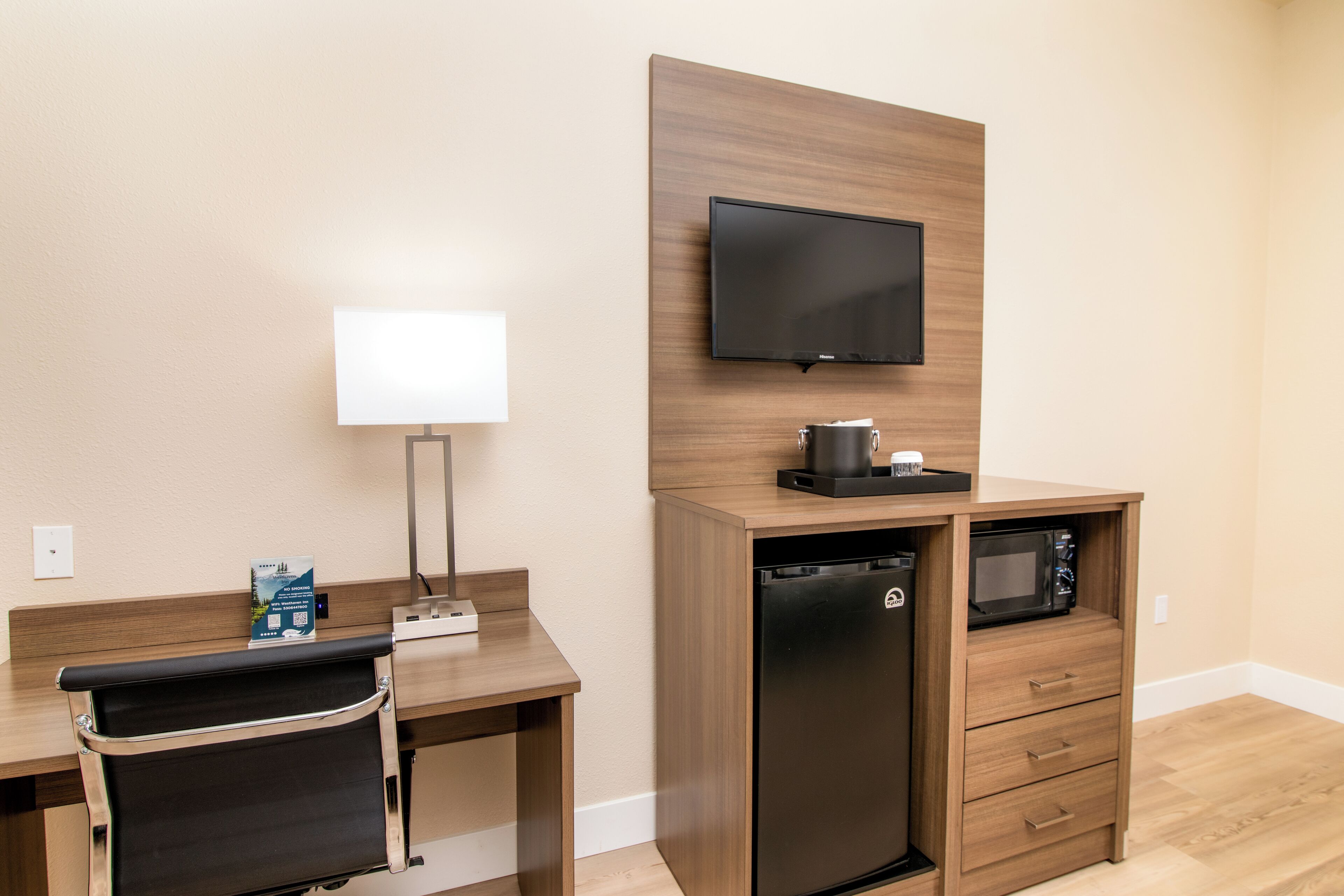 A compact hotel room with a wooden desk, a black refrigerator, a microwave, a flat-screen TV, and a small seating area with a chair.