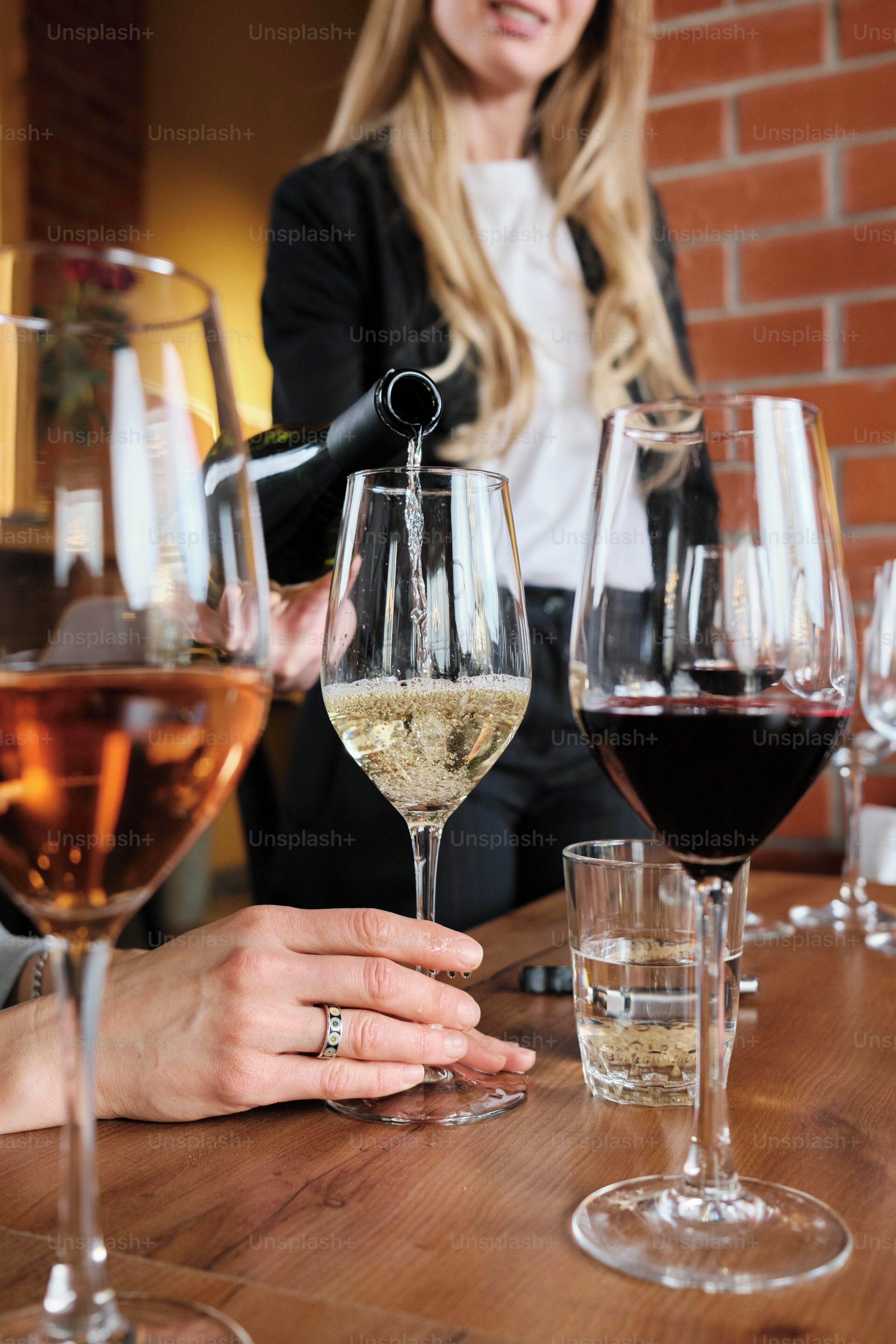 Una persona sirve una bebida en una copa de vino en un bar, con otras copas y una pared de ladrillo al fondo.