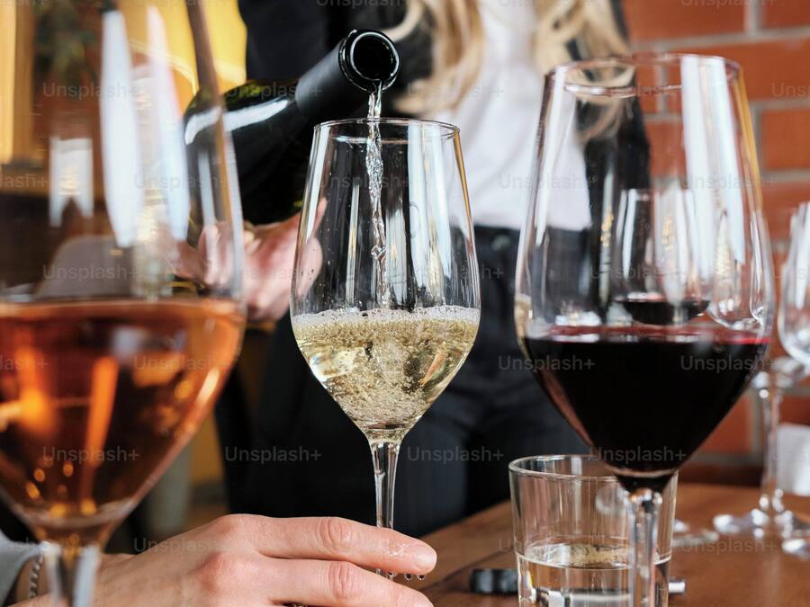 Una persona sirve una bebida en una copa de vino en un bar, con otras copas y una pared de ladrillo al fondo.