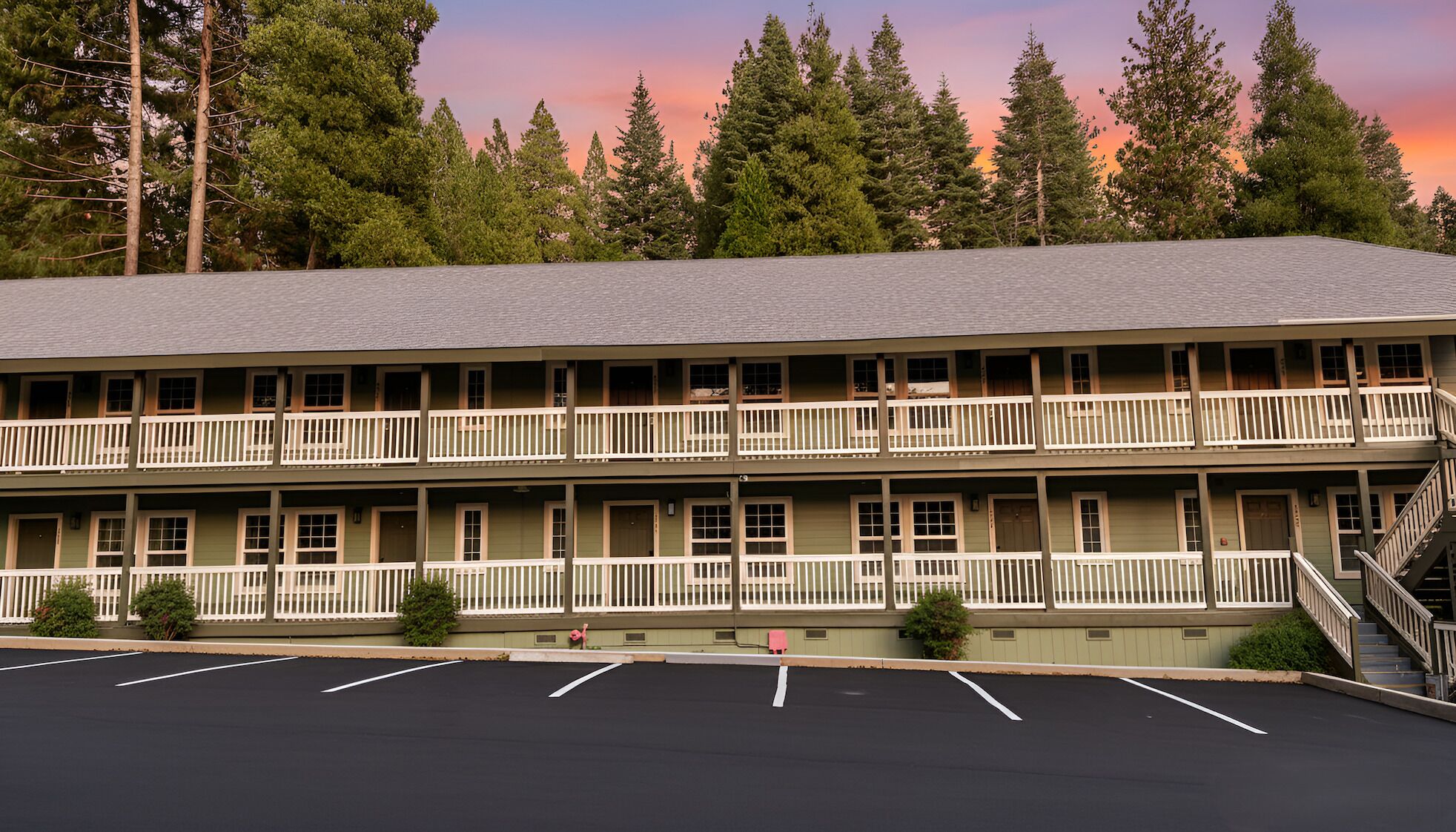 Um motel de dois andares com estacionamento em frente, cercado por árvores altas e um céu ao pôr do sol.