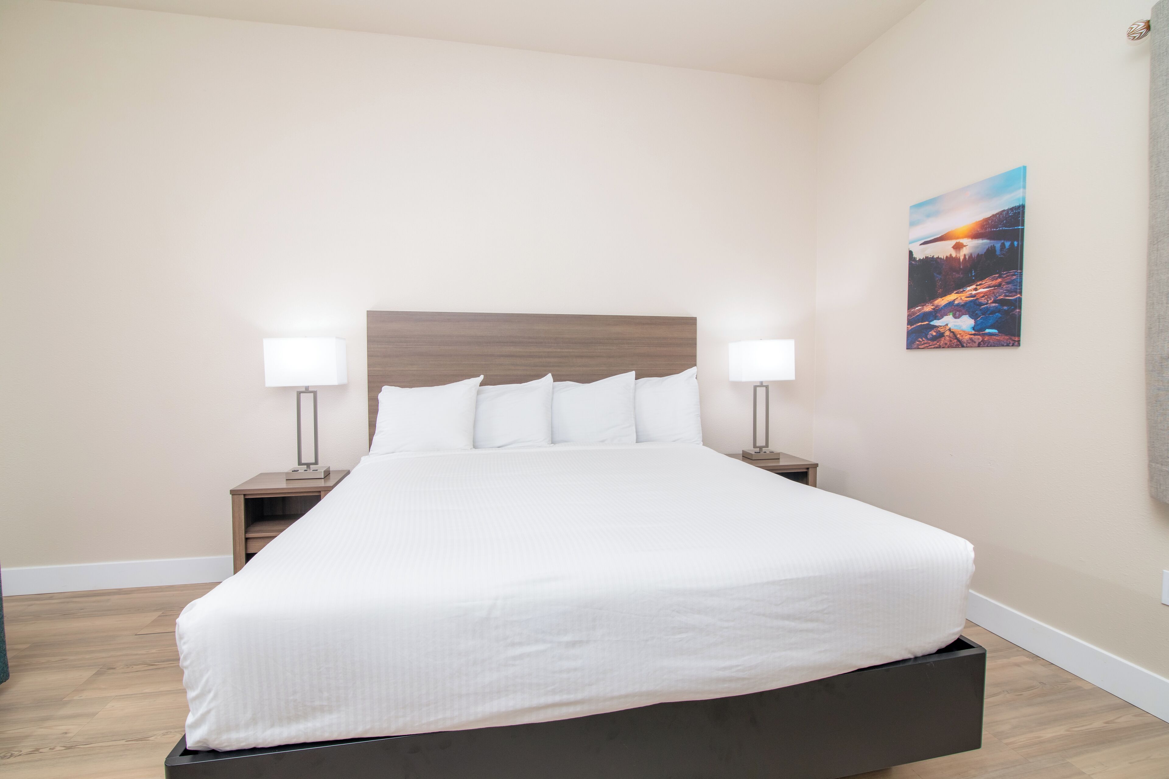 A neatly made bed with white linens in a room with light-colored walls and a wooden floor. There are two bedside tables with lamps and a framed picture on the wall.