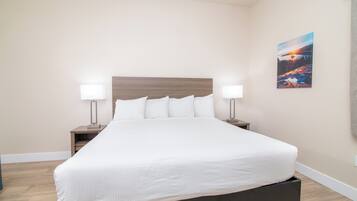 A neatly made bed with white linens in a room with light-colored walls and a wooden floor. There are two bedside tables with lamps and a framed picture on the wall.