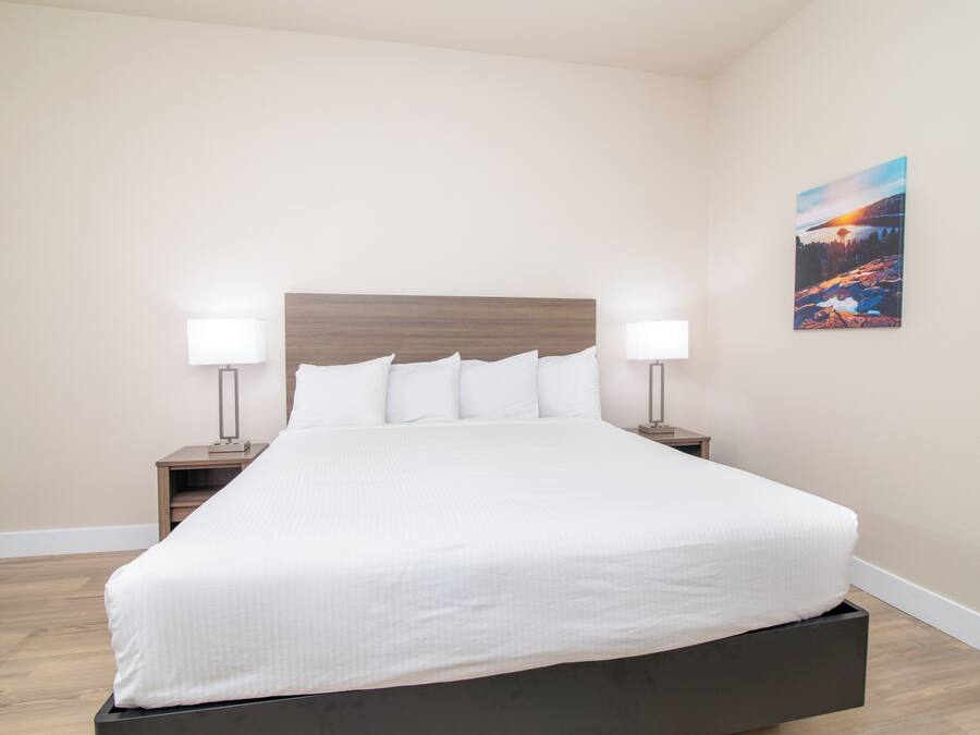 A neatly made bed with white linens in a room with light-colored walls and a wooden floor. There are two bedside tables with lamps and a framed picture on the wall.