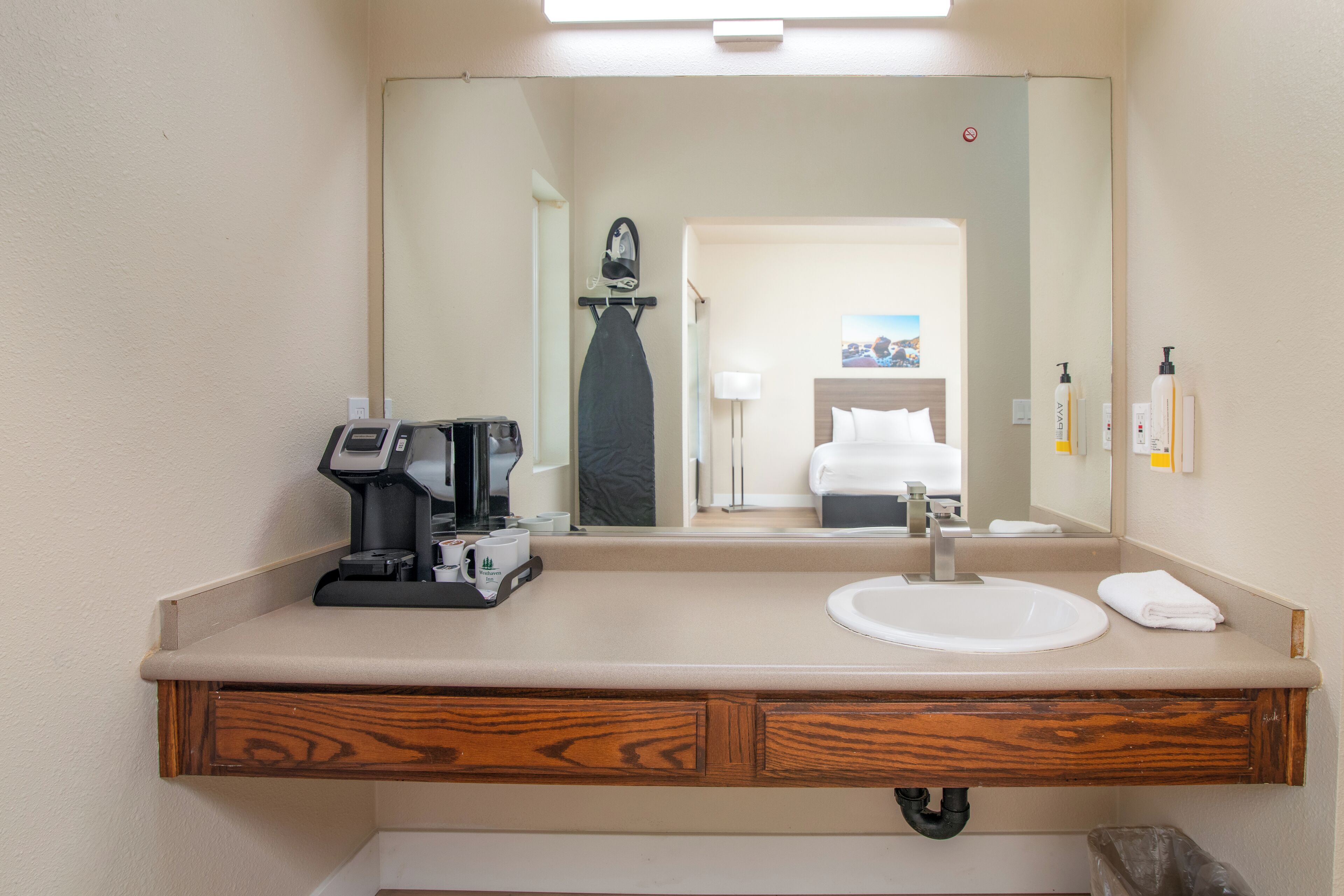 A hotel bathroom with a large mirror, a sink, a coffee maker, and a trash bin.