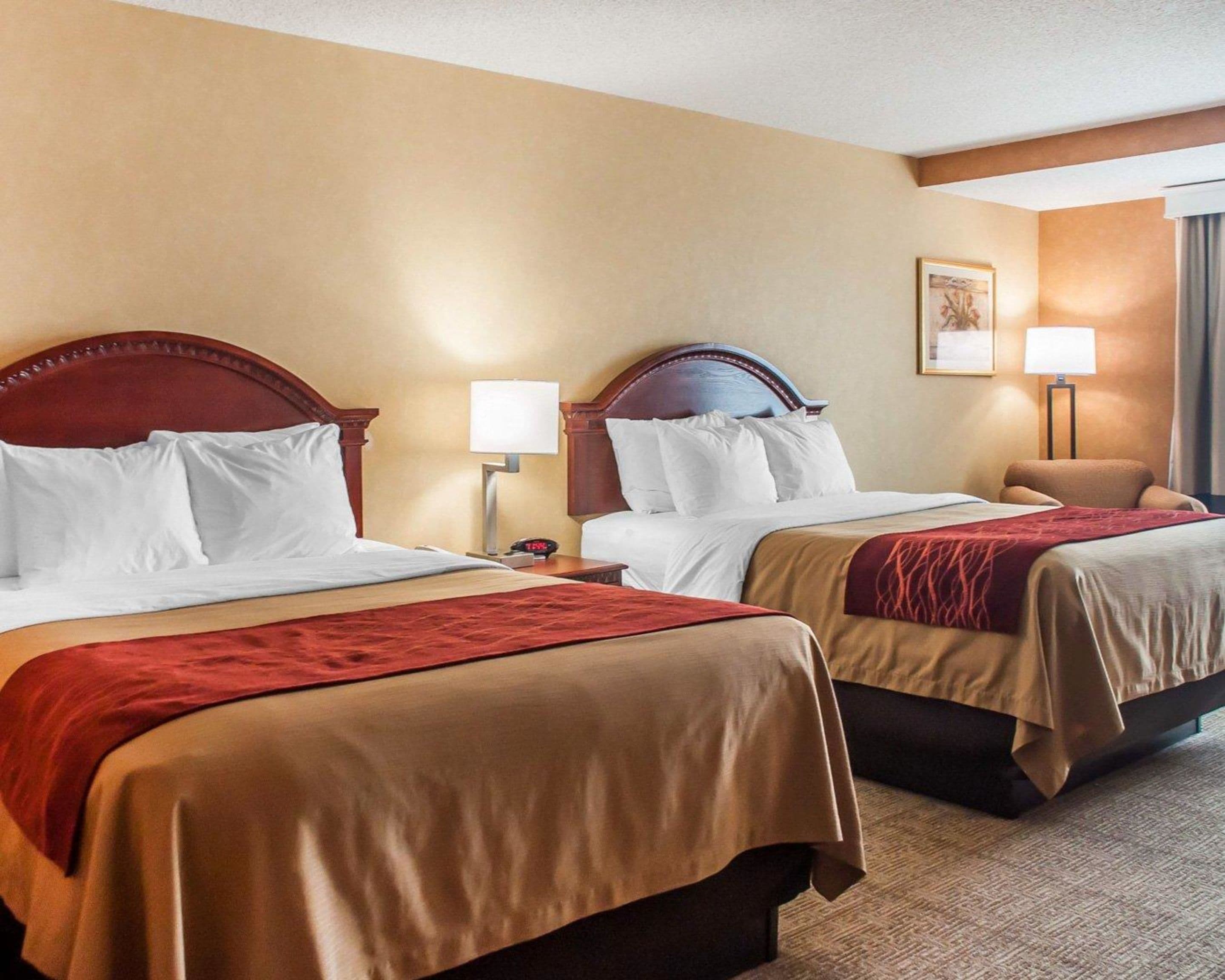 A hotel room with two beds, each with a wooden headboard, white linens, and a red throw blanket. There is a nightstand with a lamp and a clock on each bed.
