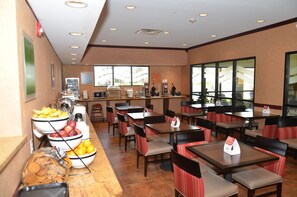 Un restaurant doté de nombreuses tables et chaises, d’un espace buffet avec des saladiers remplis de fruits et de collations, et offrant une vue sur l’extérieur grâce à de grandes fenêtres.