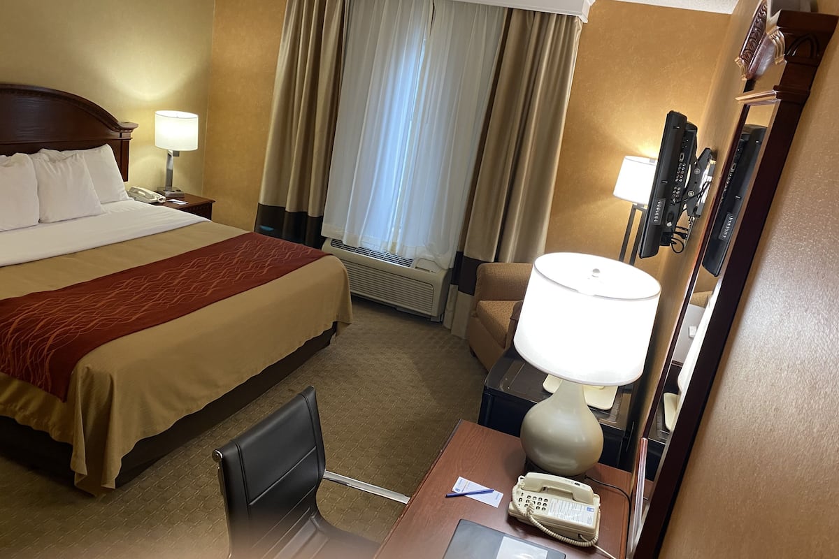 A hotel room with a bed, a desk with a telephone and a lamp, a chair, a desk lamp, and a large mirror.