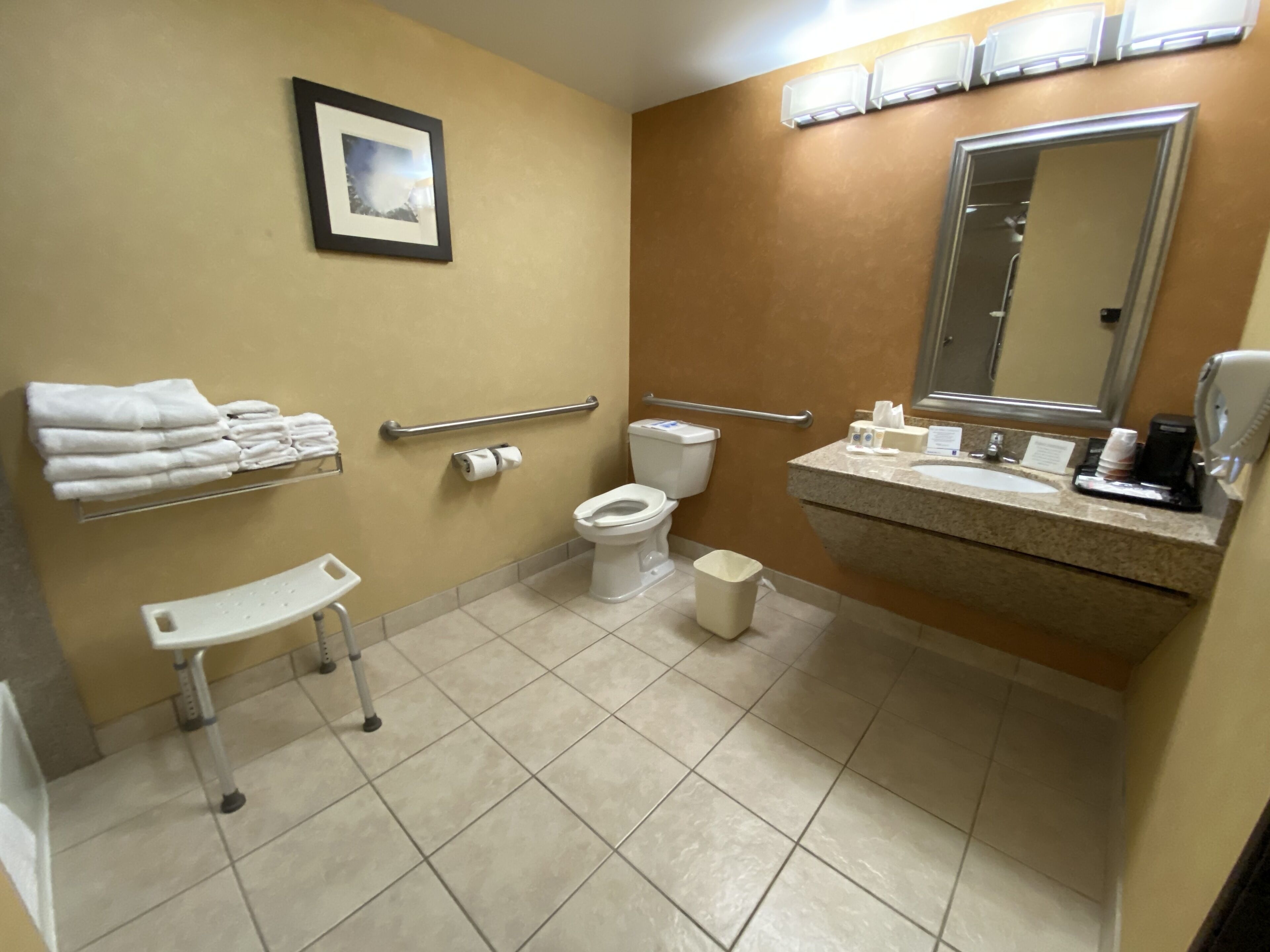 A bathroom with a toilet, sink, handrails, a trash bin, and a small white stool.