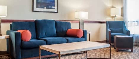 Habitación de hotel con un sofá azul, una mesa de centro de madera y una silla. Hay dos lámparas a cada lado del sofá.