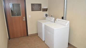 A laundry room with two white front-loading washers and dryers, a wooden door with a glass panel, and a sign above the machines.