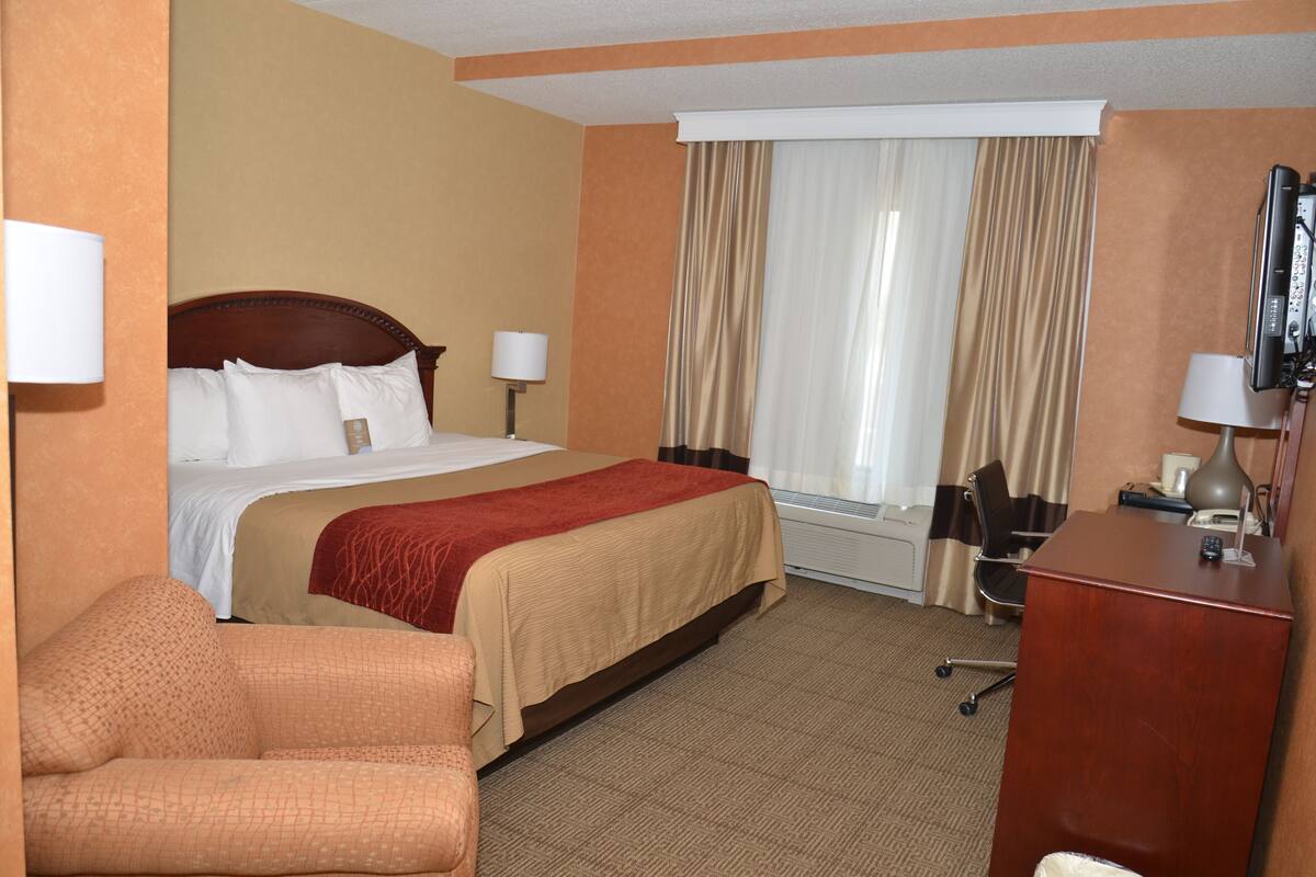 A hotel room with a large bed, two bedside lamps, a desk with a computer, a chair, and a window with curtains.