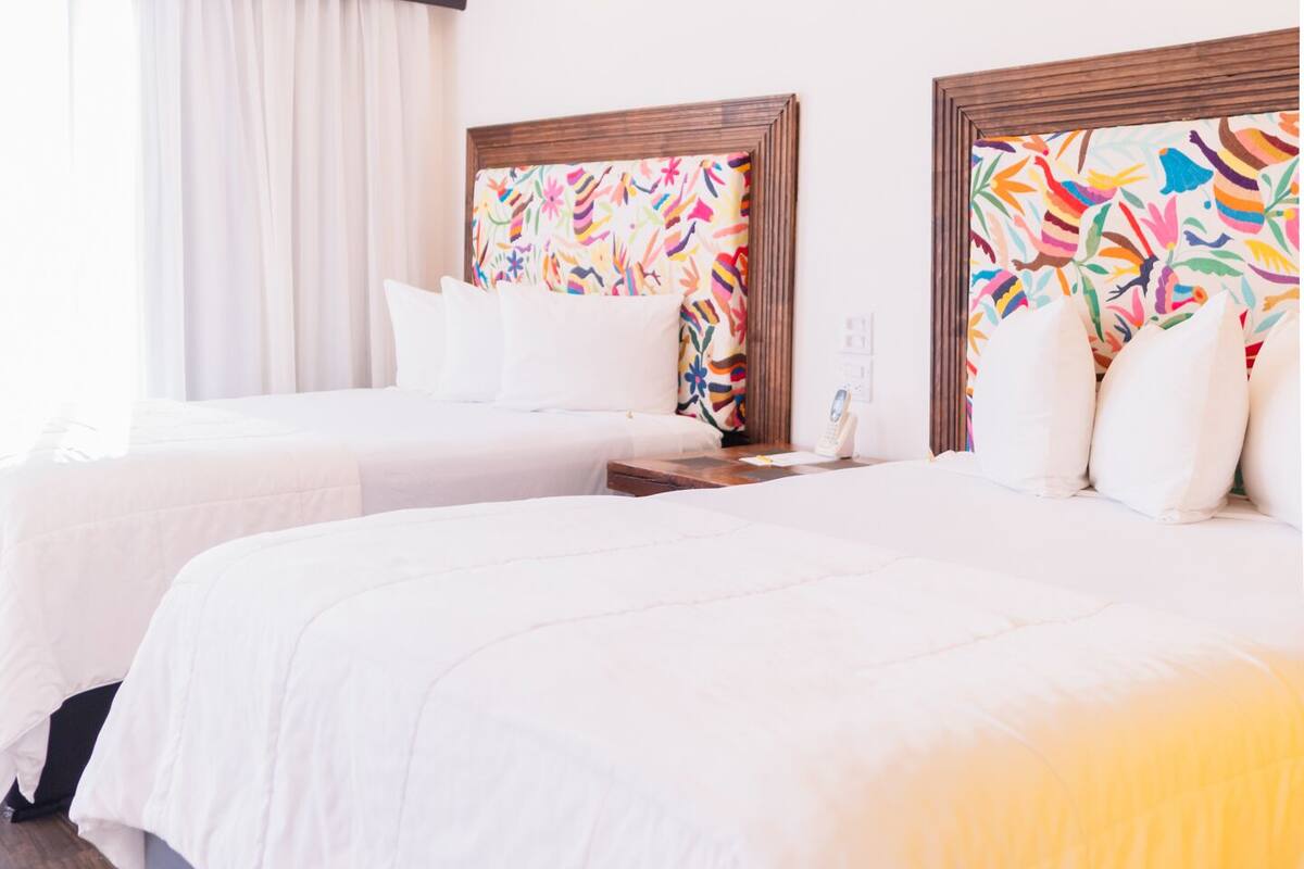 Una habitación de hotel con dos camas, ropa de cama blanca, un cabecero colorido y una ventana con cortinas.