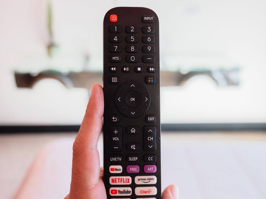 Una mano sostiene un control remoto con varios botones, incluyendo Netflix, YouTube y Plutoo.