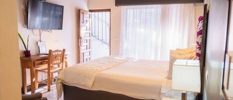 A bedroom with a bed, a desk, a chair, a TV, and a window with curtains.