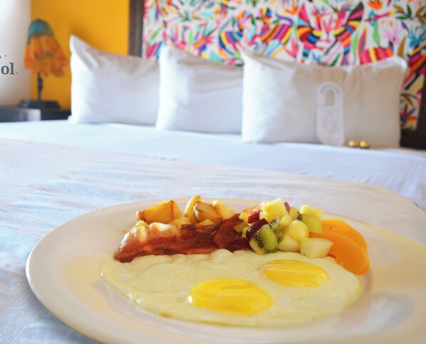 A breakfast plate with eggs, bacon, and fruit on a bed in a hotel room.