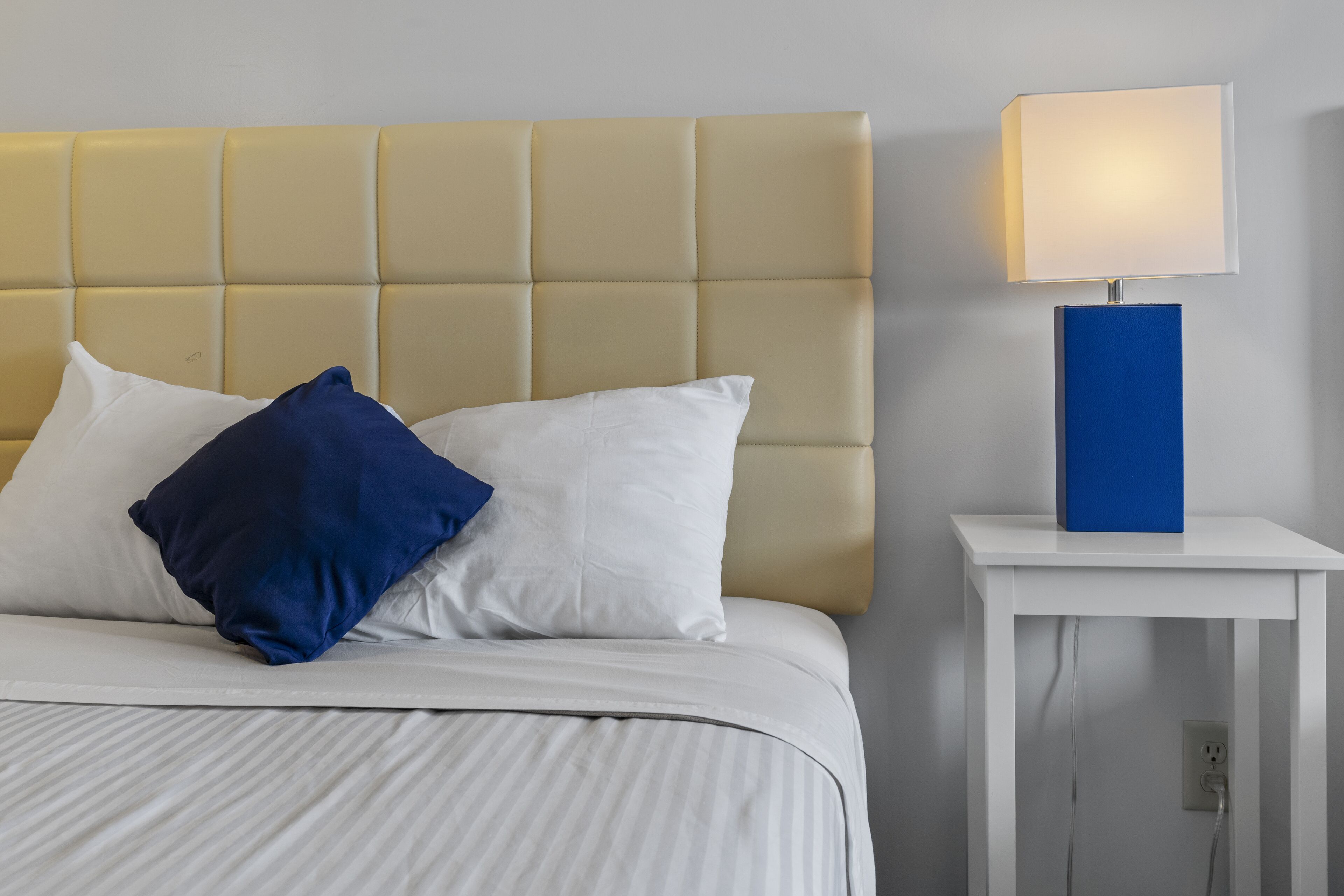 A bed with a headboard, white bedding, and two pillows (one white, one blue). A bedside table with a lamp and a power outlet is visible.