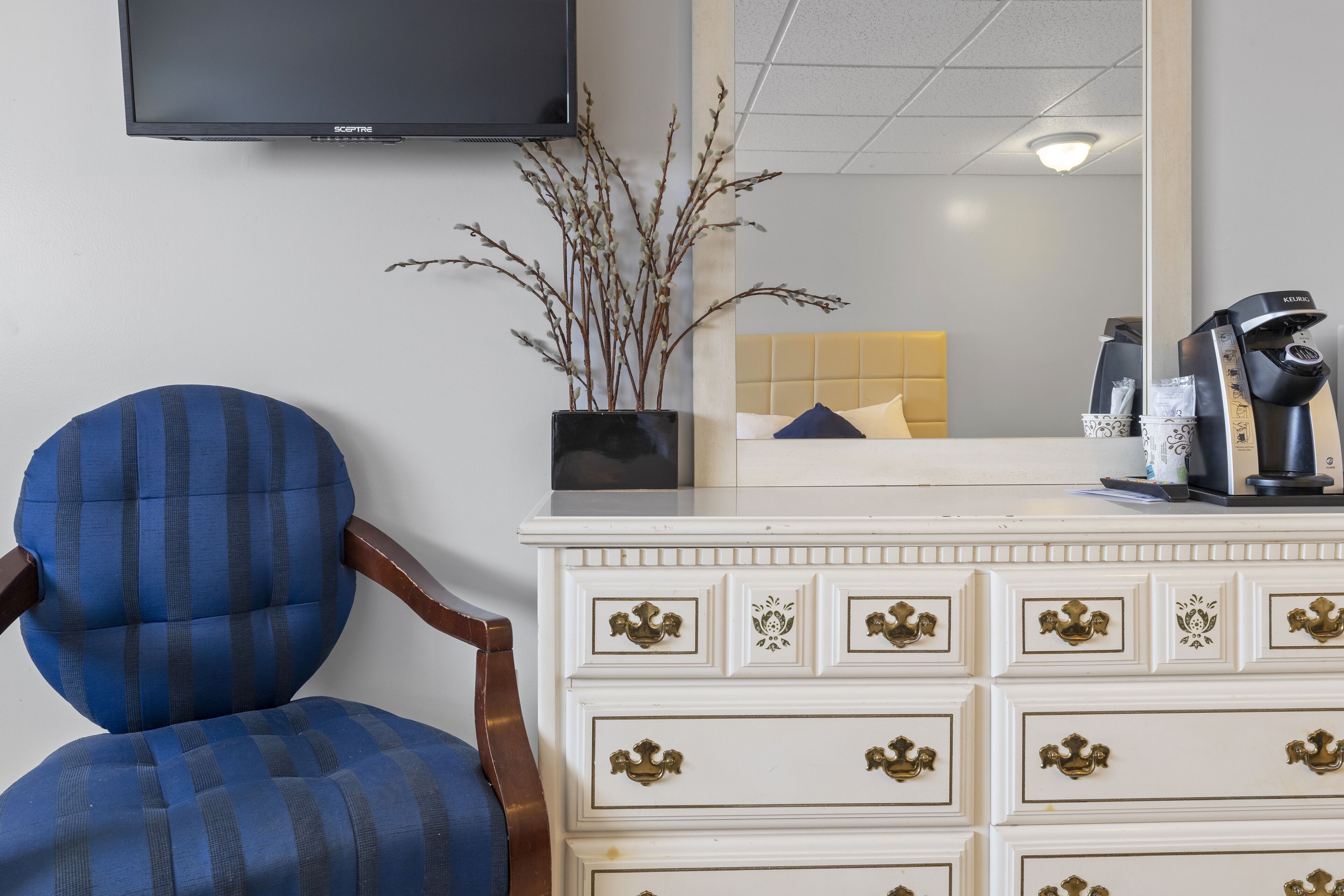 Une pièce comprenant un fauteuil recouvert d’un tissu bleu, une commode blanche avec des poignées dorées, un téléviseur à écran plat et une machine à café.