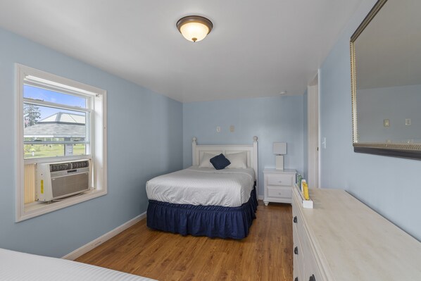 Un dormitorio con cama, ventana con aire acondicionado, mesita de noche, cómoda y espejo.