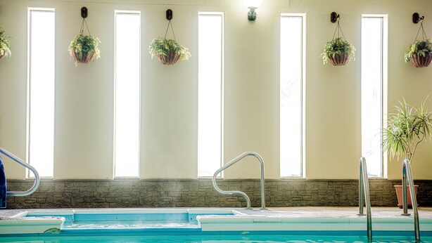 An indoor swimming pool with clear glass walls, a stone wall, and hanging planters. - Wrangler Inn  (Mobridge)