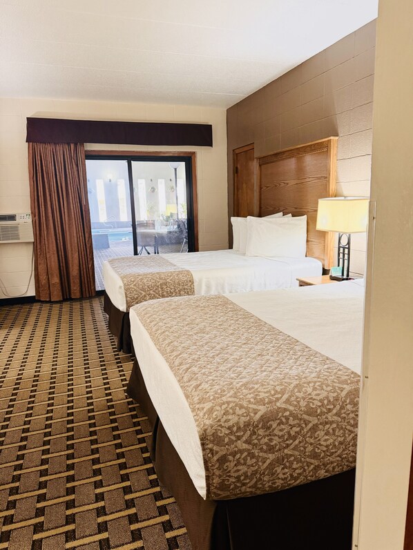 Habitación de hotel con dos camas, un ventanal con cortinas, vistas a la piscina y una mesita de noche con lámpara.