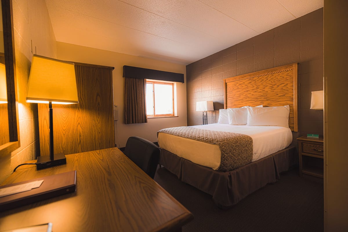 A hotel room with a wooden desk, a bed with a headboard, a nightstand, a lamp, and a window with curtains.
