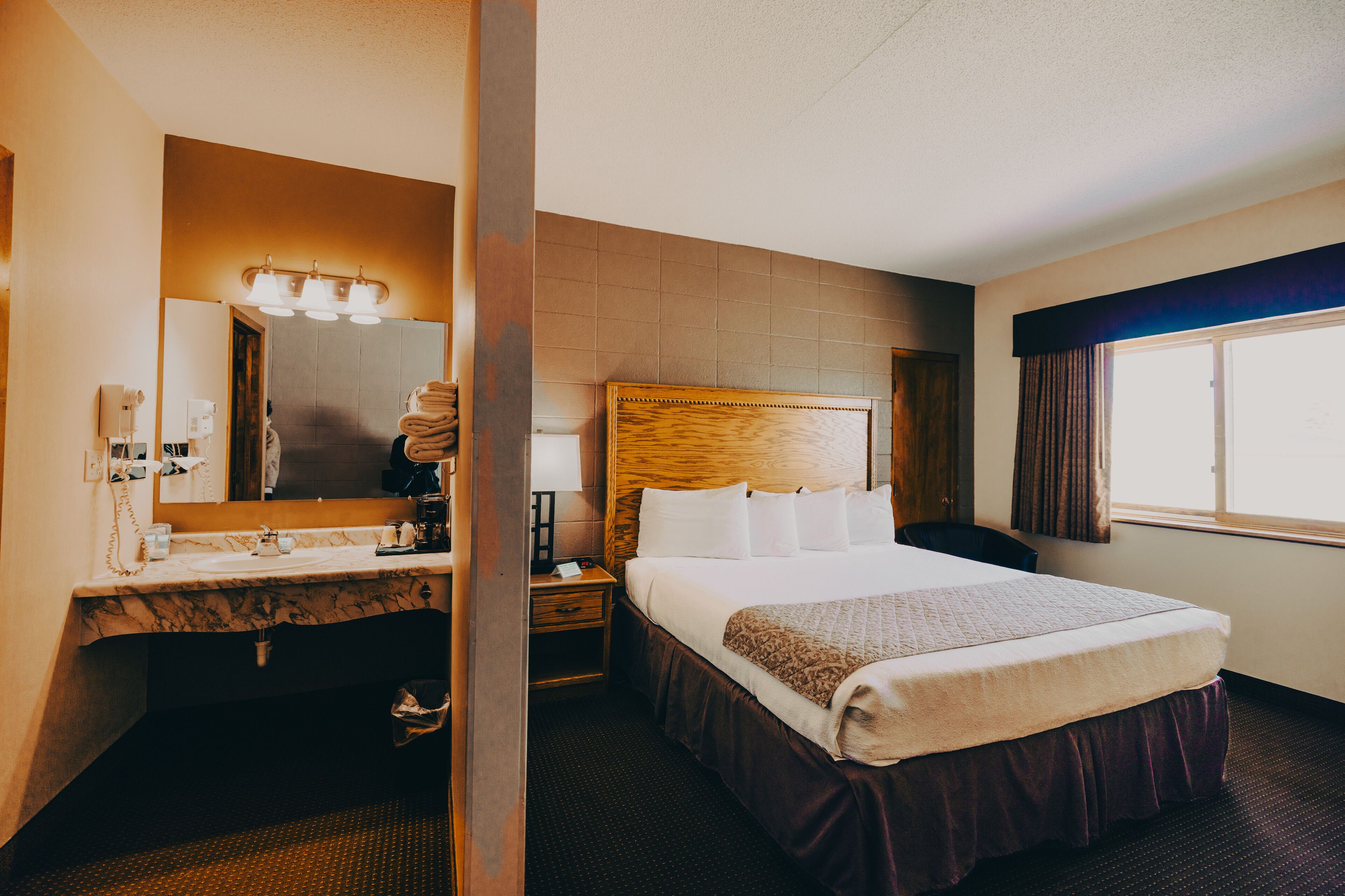 A hotel room with a large bed, a wooden headboard, a vanity with a mirror, and a window with curtains.