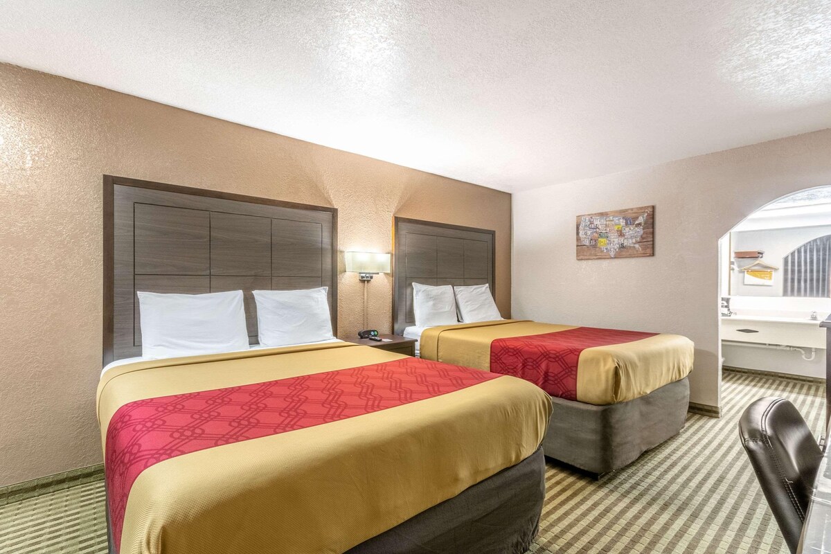 A hotel room with two beds, a desk, a chair, and a bathroom visible through an archway.