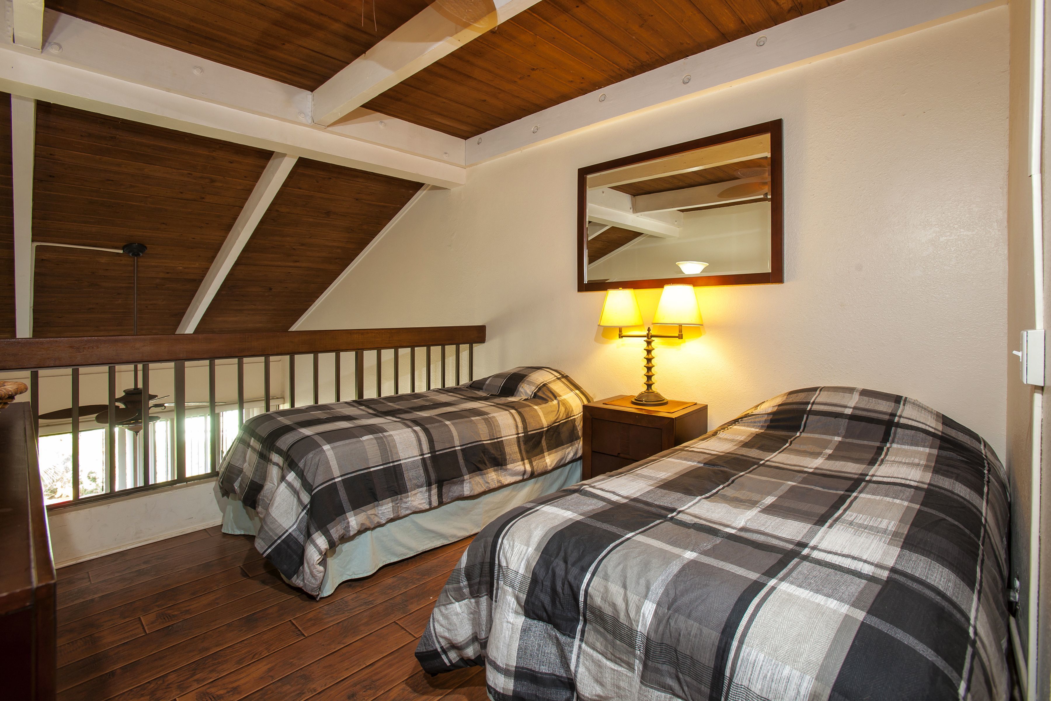 A bedroom with two beds, a wooden ceiling, a window with a view, and a nightstand with a lamp.