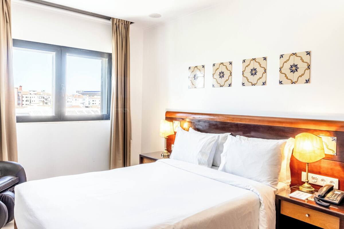 A hotel room with a large bed, a nightstand with a lamp, a chair, and a window with curtains.